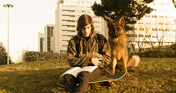 Teen sitting reading outdoors with an alsation dog and skateboard.