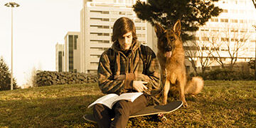 Teenager sitting on grass with dog, reading a book.